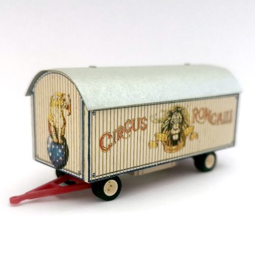 Ein Miniatur-Zirkusanhänger mit farbenfroher Grafik mit einem Löwen und dem Wort „Circus Roncalli“ auf der Seite. Der Anhänger hat ein hellblaues Dach, weiße Räder und eine rote Anhängerkupplung.