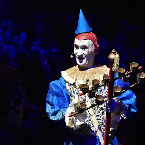 Ein Clown in blauem Kostüm und Hut tritt unter blauem Licht auf und hält einen mit Glöckchen geschmückten Stab in der Hand. Im Hintergrund ist ein Publikum zu sehen, das der Vorstellung zusieht.