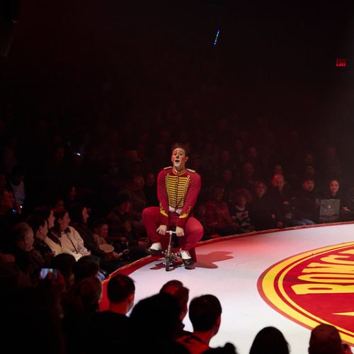 Ein Zirkusartist in einem rot-gelben Kostüm sitzt auf einem kleinen Hocker in der Mitte einer Manege, umgeben von einem großen Publikum. Die Manege ist hell beleuchtet und die Menge schaut in der schwach beleuchteten Arena gespannt zu.