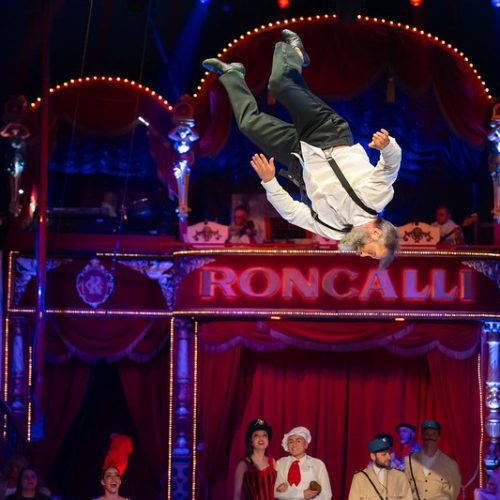 Ein Artist in weißem Hemd und schwarzer Hose wirbelt unter hellem Zirkuslicht durch die Luft. Im Hintergrund ist eine rot-goldene Zirkuskulisse mit einem zuschauenden Publikum zu sehen. Auf dem Schild steht „Roncalli“.