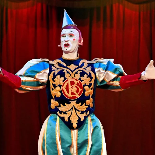 Ein als Clown verkleideter Darsteller steht mit weit ausgebreiteten Armen auf der Bühne. Das Kostüm ist farbenfroh und in Blau, Grün, Rot und Gold gehalten und trägt ein dekoratives Emblem auf der Brust. Der Hintergrund ist ein tiefroter Vorhang.