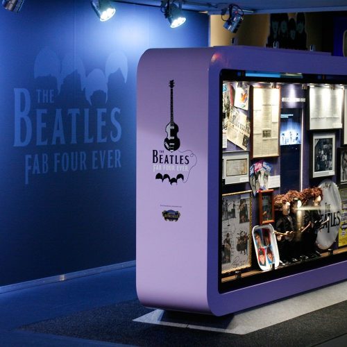 Ausstellungsstück in einem Museum mit Beatles-Erinnerungsstücken. Der Bereich ist blau beleuchtet und verfügt über ein auffälliges Schild mit der Aufschrift „The Beatles Fab Four Ever“. Die Vitrine enthält Fotos, Instrumente und verschiedene bandbezogene Gegenstände.