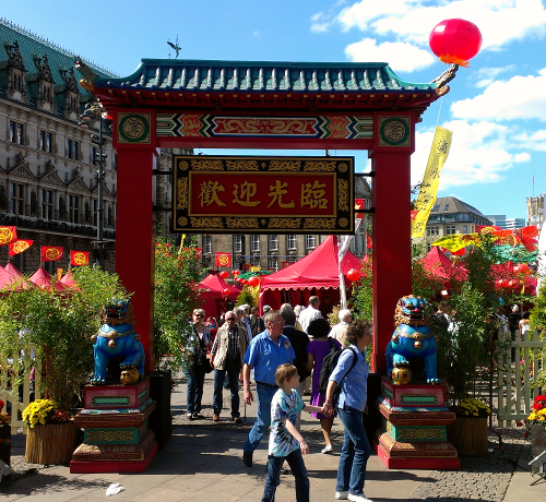 Menschen gehen bei einem lebhaften Open-Air-Festival durch ein dekoratives chinesisches Tor. Man sieht rote Laternen und Zelte, im Hintergrund sieht man Grün und ein historisches Gebäude. Die Szene ist lebhaft und farbenfroh, unter einem sonnigen Himmel.