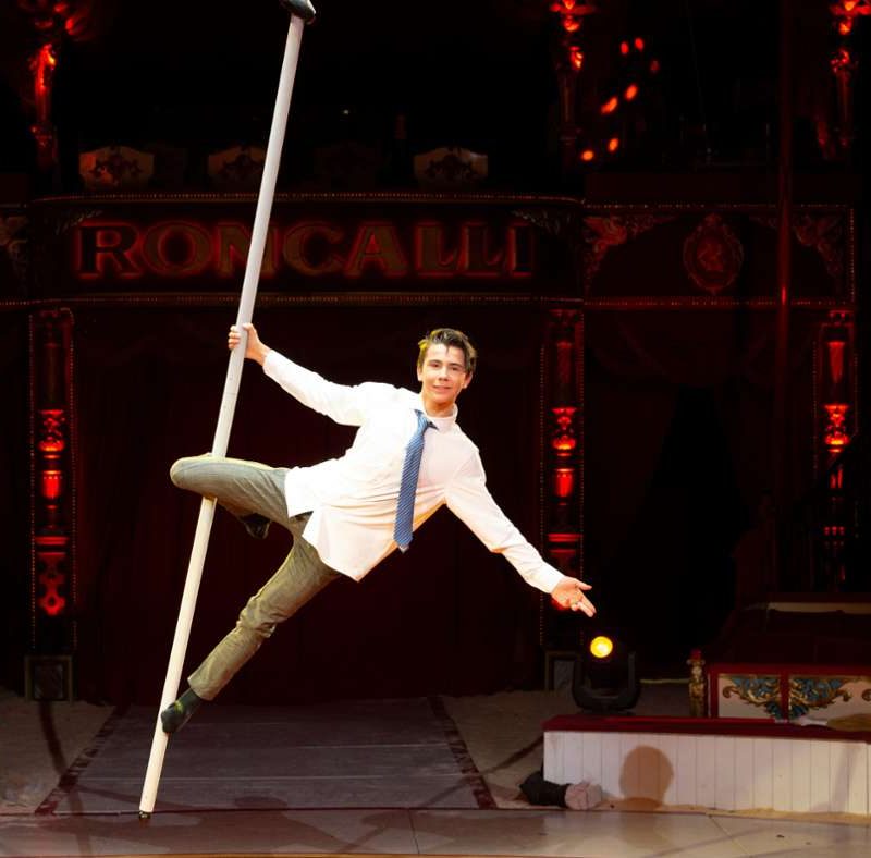 Ein Artist in weißem Hemd und Krawatte balanciert gekonnt seitlich auf einer vertikalen Stange in einer Zirkuskulisse, mit rot-goldenem Dekor und dem Wort „RONCALLI“ im Hintergrund. Bühnenlichter erhellen die Szene.