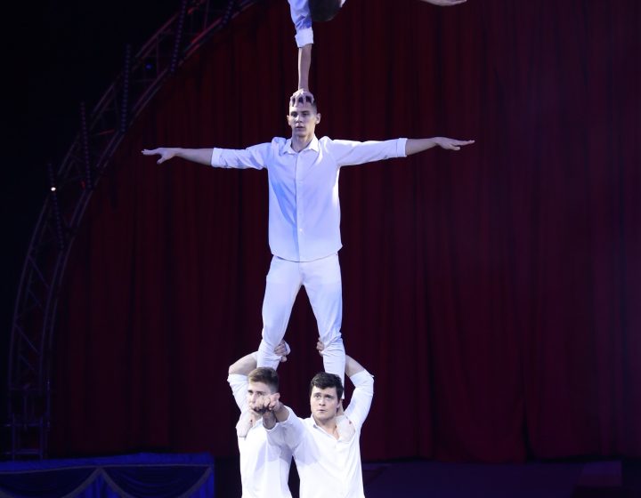 Vier Akrobaten in weißen Kostümen führen auf der Bühne eine menschliche Pyramide auf, wobei einer kopfüber an der Spitze balanciert. Ein runder Rahmen und ein roter Vorhang bilden den Hintergrund unter dem Scheinwerferlicht.