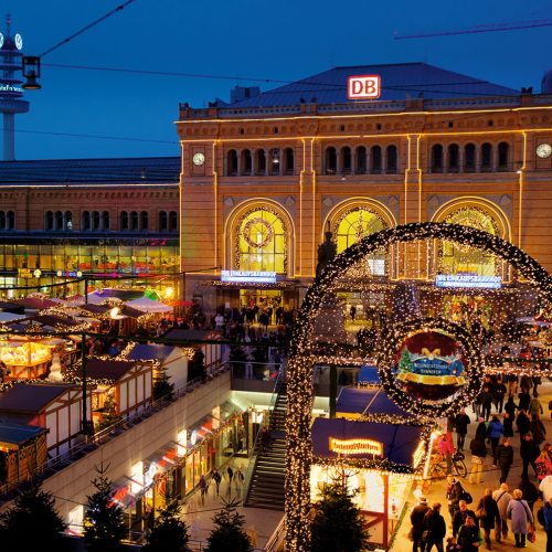 Ein lebhafter Weihnachtsmarkt bei Nacht vor einem hell erleuchteten Bahnhof mit „DB“-Beschriftung. Die Stände sind mit Lichtern und Dekorationen geschmückt, während Scharen von Menschen durch die festliche Szene unter einem beleuchteten Torbogen schlendern.