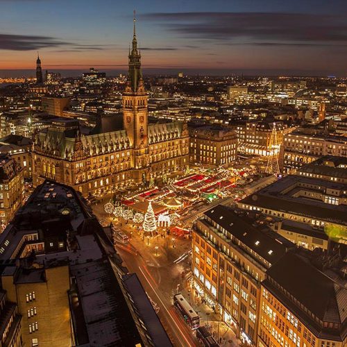 Luftaufnahme eines Stadtplatzes in der Abenddämmerung mit einem hell erleuchteten Weihnachtsmarkt mit verschiedenen Ständen und Dekorationen. Das historische Rathaus mit seinem beleuchteten Glockenturm sticht zwischen den umliegenden Gebäuden und den Lichtern der Stadt hervor.