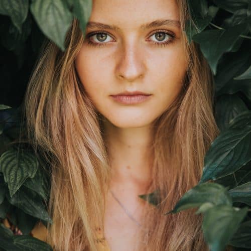 Eine Frau mit langen blonden Haaren blickt direkt in die Kamera, teilweise eingerahmt von üppigen grünen Blättern. Sie trägt ein gelbes Blumentop und das natürliche Licht beleuchtet sanft ihr Gesicht.