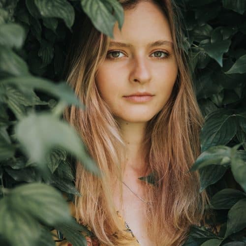 Eine Frau mit langem blondem Haar steht inmitten dichtem grünen Laub. Ihr Gesichtsausdruck ist ruhig, und sie trägt ein gelbes Oberteil mit Blumenmuster. Die Blätter rahmen teilweise ihr Gesicht ein und schaffen eine natürliche, ruhige Atmosphäre.
