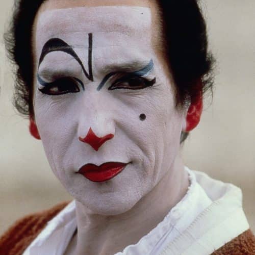 Eine Person mit dramatischer weißer Gesichtsbemalung, kräftigem schwarz-rotem Make-up und einem aufgemalten Schönheitsfleck, die eine rote Strickjacke über einem weißen Hemd trägt, blickt mit ernstem Gesichtsausdruck leicht zur Seite.