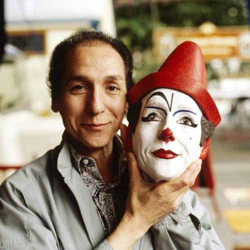 Ein Mann lächelt und hält eine bemalte Clownsmaske mit einem roten Hut neben sein Gesicht, während er draußen in einem Café oder auf einem Markt steht.