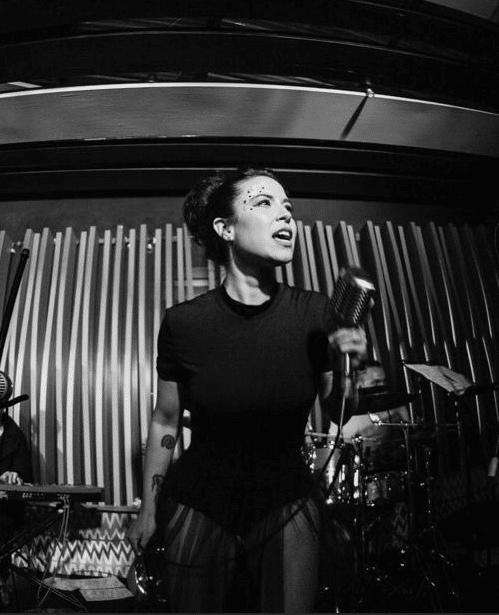 Eine Frau in einem figurbetonten Outfit singt auf der Bühne in ein Vintage-Mikrofon. Hinter ihr befinden sich dramatische Beleuchtung und vertikale Wandpaneele. Im Hintergrund sind teilweise ein Schlagzeug und ein Keyboarder zu sehen.